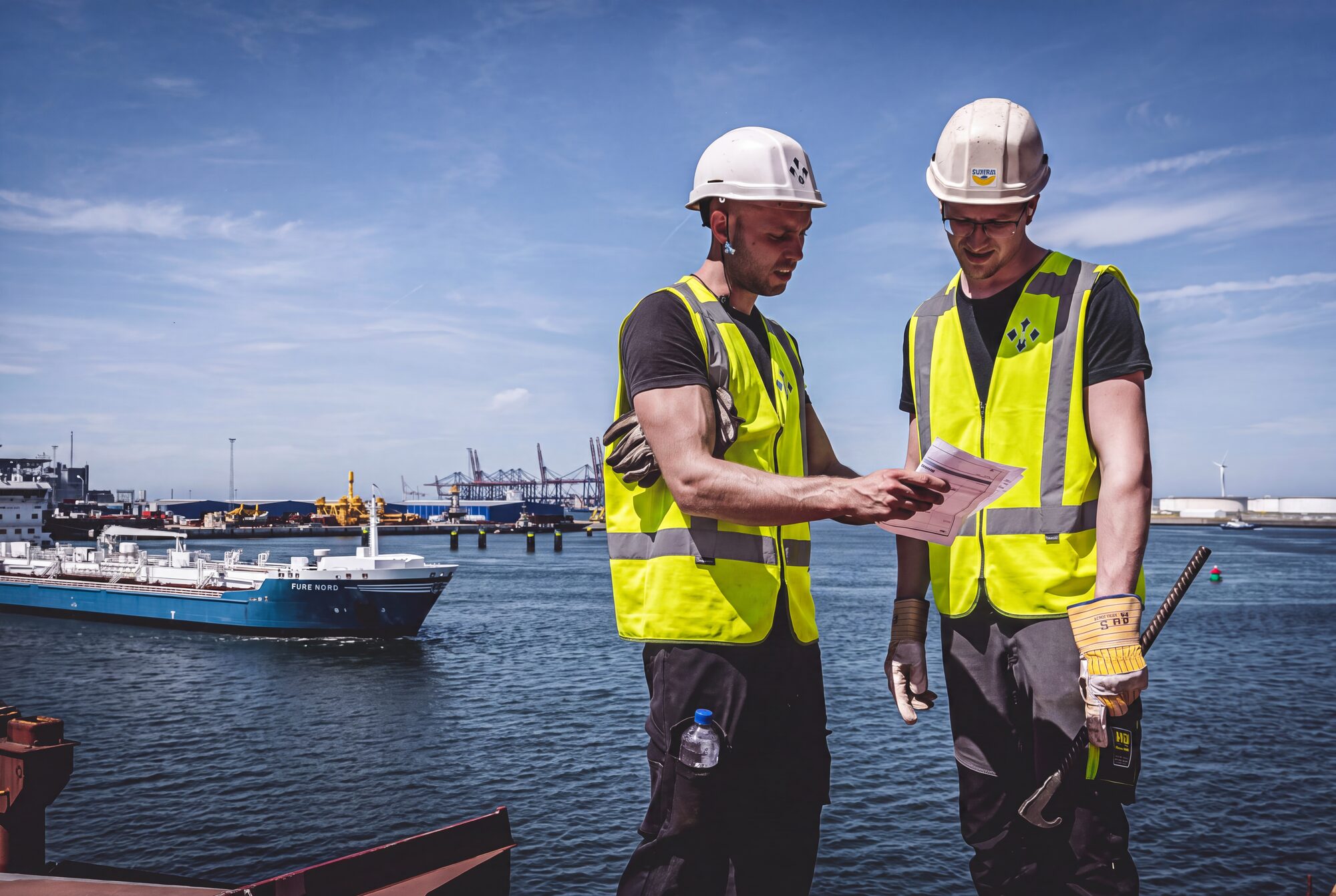 Twee havenwerkers in geel hesje overleggen over een tekening in de Rotterdamse haven